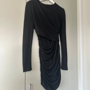 Svart klänning från Zara - Elegant svart klänning från Zara med draperad design och åtsittande passform. Klänningen har långa ärmar och vaddering i axlarna med en öppning på sidan av överdelen. Perfekt för fest eller middag! Endast använd 1 gång 