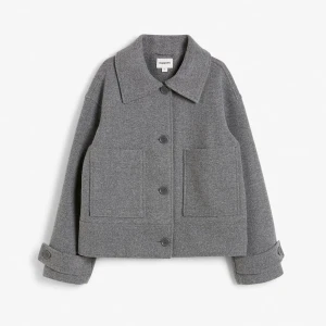 Grå kappa från Kappahl  - Snygg grå kappa från Kappahl med stora fickor och knappar framtill. Jackan har en klassisk krage och långa ärmar med knappar vid ärmsluten. Perfekt för en stilren look.