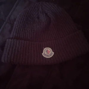 Svart ribbad mössa från Moncler - Snygg svart ribbad mössa från Moncler med uppvikt kant och logotyp framtill. Perfekt för att hålla värmen under kyliga dagar.