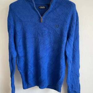 Blå tröja med dragkedja från Chaps - Säljer en snygg blå tröja från Chaps i bomull. Tröjan har en dragkedja vid halsen och ett diskret broderat emblem på bröstet. Perfekt för en stilren look.