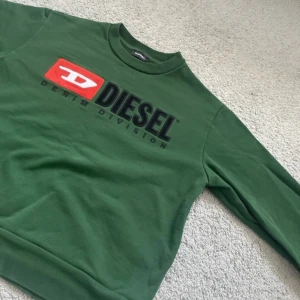 Grön sweatshirt från Diesel - Säljer en grön sweatshirt från Diesel med ett stort tryck på framsidan. Tröjan har långa ärmar och en rund halsringning. Perfekt för en avslappnad stil. 