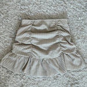 Beige rutig kjol från Gina Tricot - Beige rutig kjol från Gina Tricot i storlek XS. Kjolen har en volangdetalj med rynkade partier. 