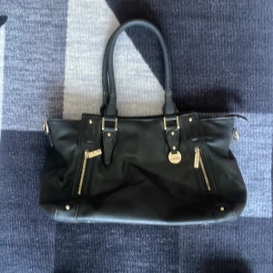 Svart axelväska med dragkedjor - Snygg svart axelväska i skinn med praktiska dragkedjor och guldfärgade detaljer. Perfekt för att bära med sig allt man behöver under dagen. Väskan har två robusta handtag och en stilren design.