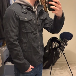 Svart jacka med dragkedja och knappar - Snygg svart jacka med dragkedja och knappar. Den har flera fickor framtill och en huva i grått tyg. Perfekt för en casual look. Jackan är i väldigt bra skick, perfekt till våren. Nypris på denna är runt 1300kr. Skriv vid funderingar! 