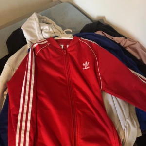 Oanvänd Röd träningsjacka från Adidas - Snygg röd träningsjacka från Adidas med klassiska vita ränder längs ärmarna. Jackan har dragkedja framtill och Adidas-logga på bröstet. Perfekt för en sportig look eller träning.