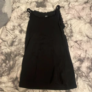 Svart klänning från H&M - Snygg svart klänning från H&M med spetsdetaljer vid axelbanden. Perfekt för en stilren look. Klänningen är ärmlös och har en enkel design som passar till många tillfällen.