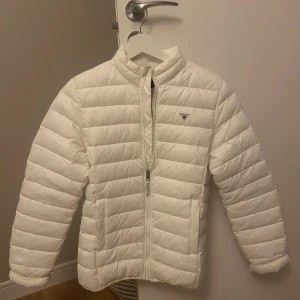 Vit dunjacka från Gant - Säljer en stilren vit dunjacka från Gant, helt oanvänd. Jackan har en dragkedja framtill och är långärmad med en normal passform. 