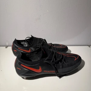 Nike Phantom fotbollsskor - Säljer ett par svarta Nike Phantom fotbollsskor med röda detaljer. Skorna har snörning och är designade för optimal prestanda på planen. Perfekta för dig som vill ha stil och funktion i ett.