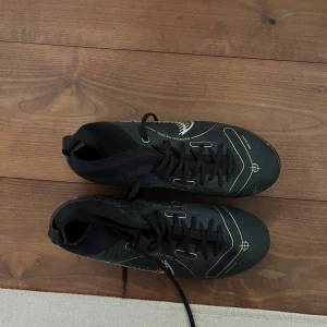 Svarta fotbollsskor från Nike - Snygga svarta fotbollsskor från Nike med vita och guldiga detaljer. Skorna har snörning och en hög ankelkrage för extra stöd. Perfekta för fotbollsplanen med sina dobbar för bra grepp, gjorde för gräs. Endast använd 1-2 gånger