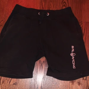 Svarta shorts från Sail Racing passar bra till sommaren  - Snygga svarta shorts från Sail Racing med logga på benet. De har en bekväm passform med snörning i midjan och en bakficka. Perfekta för en avslappnad stil.