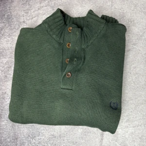 Grön tröja från Chaps - Säljer en grön tröja från Chaps i storlek XL. Tröjan är tillverkad av bomull och har en ribbad krage med knappar. Perfekt för kyligare dagar med sin långärmade design och diskreta logga på bröstet.