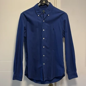 Blå skjorta från Ralph Lauren - Snygg blå skjorta från Ralph Lauren i slim fit. Skjortan har långa ärmar och knappar framtill samt på kragen. Perfekt för en stilren look.