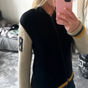 Marinblå och beige stickad kofta med dragkedja - Säljer en snygg marinblå och beige stickad kofta med dragkedja. Koftan har långa ärmar med ett nummer på ärmen och gula detaljer vid ärmslut och nederkant. Perfekt för en avslappnad stil.