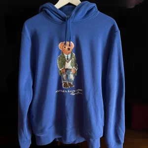 Blå hoodie från Ralph Lauren - Säljer en snygg blå hoodie från Ralph Lauren med en ikonisk nallebjörn på framsidan i stl M. Hoodien är sparsamt använd och i mycket fint skick. Ordinarie pris ca 2000.