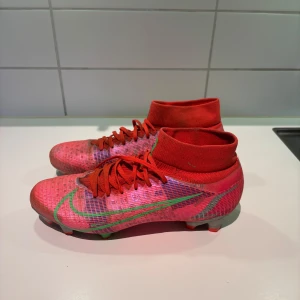 Nike Mercurial Superfly 8 PRO, fotbollssko - Nike Mercurial Superfly 8 PRO FG. Snygga röd/Rosa fotbollsskor från Nike med grön och silverdetaljer. Skorna har snörning och en strumpliknande passform för extra stöd. Perfekta för fotbollsplanen med sin greppvänliga sula. Skorna är använda men absolut inte trasig så ett bra skick för ett par fina fotbollsskor 