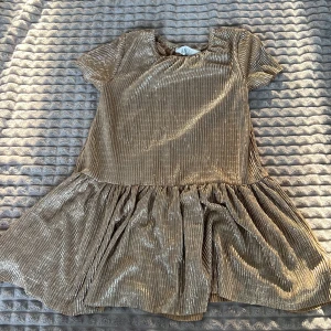 Guldglittrig klänning - Säljer en glittrig guldklänning med korta ärmar och volangdetaljer. Perfekt för festliga tillfällen. Klänningen har en lös passform och är tillverkad i ett skimrande material som fångar ljuset vackert.