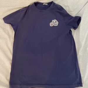 Blå t-shirt från Moncler - Säljer en snygg blå t-shirt från Moncler med ett coolt broderat märke på bröstet. Perfekt för en avslappnad stil. Passar bra till jeans eller shorts!