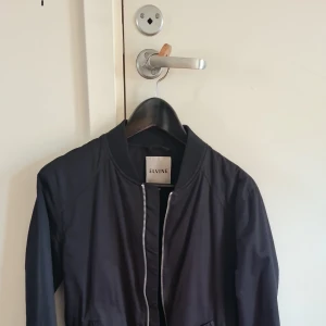 Svart bomberjacka från Elvine - Snygg svart bomberjacka från Elvine i storlek XS. Jackan har en dragkedja framtill och ribbade muddar vid ärmslut och nederkant. Perfekt för en stilren look.