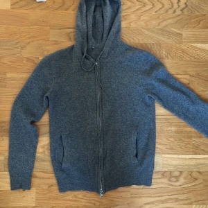Grå hoodie med dragkedja - Säljer en stilren grå hoodie med dragkedja och huva. Tröjan har långa ärmar och ribbade muddar vid ärmslut och nederkant. Perfekt för kyliga dagar.