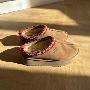 UGG TAZZ Tofflor - Säljer ett par bruna mockaskor från UGG med röd och vit kantdetalj. Skorna har en mjuk insida och en robust sula, perfekt för kalla dagar. De har en enkel design med en liten logga på sidan.