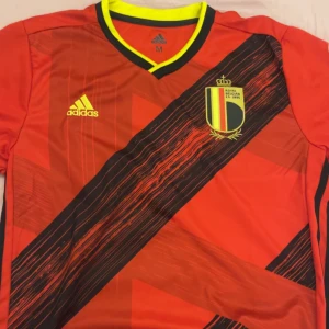 Röd Belgien fotbollströja från Adidas - Snygg röd Belgien fotbollströja från Adidas med svart och gult mönster. Tröjan har korta ärmar och en V-ringad krage med gula detaljer. Perfekt för fotbollsfans som vill visa sitt stöd under UEFA Euro 2020.☀️☀️☀️