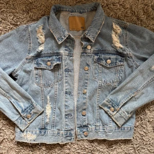 Blå jeansjacka med slitningar - Säljer en fin blå jeansjacka från Lager157 med slitna detaljer för en sliten look. Jackan har knappar framtill och två bröstfickor med lock. Normal passform! Perfekt till sommaren☀️🩵