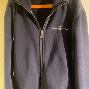 Mörkblå hoodie från Sail Racing - Säljer en snygg mörkblå hoodie från Sail Racing med dragkedja och justerbar luva. Storlek L och i väldigt bra skick. 👍 Priset inte hugget i stenen