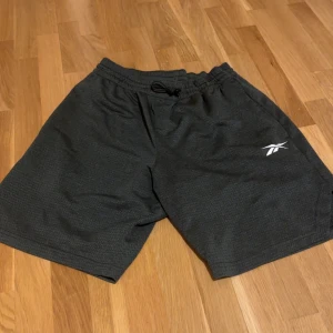 Svarta träningsshorts från Reebok - Snygga svarta träningsshorts från Reebok med elastisk midja och snörning. Perfekta för gymmet eller löprundan. Diskret logga på ena benet.
