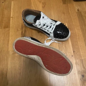 Louboutins !Söker byte! - Unika sneakers i svart och vitt med nitar på tån och sidorna. Skorna har en röd sula och vita snören. Perfekta för att ge en edgy touch till din outfit.
