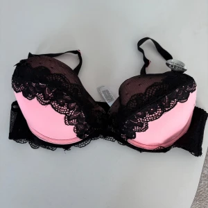 Rosa och svart push-up BH - Snygg rosa och svart push-up BH med spetsdetaljer och avtagbara inlägg. Den har justerbara axelband och en dekorativ rosett framtill. Perfekt för att ge extra lyft och form. Oanvänd. 