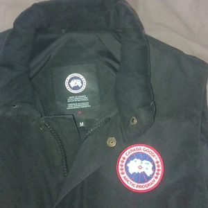 Svart dunväst från Canada Goose - Säljer en svart dunväst från Canada Goose med det klassiska märket på bröstet. Västen har knappar och dragkedja framtill för extra skydd mot kyla. Perfekt för kyliga dagar.