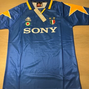 Blå fotbollströja från Kappa - Säljer en klassisk blå fotbollströja från Kappa med gula detaljer och korta ärmar. Tröjan har ett tryck med 'SONY' på framsidan och 'ZIDANE 21' på baksidan. Perfekt för fotbollsfans som älskar retrostil! ⭐️