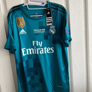Blå Real Madrid fotbollströja - Säljer en blå Real Madrid fotbollströja från Adidas med 'Fly Emirates' tryck på framsidan. Tröjan har korta ärmar och ett FIFA-märke på bröstet. På baksidan finns namnet 'Ronaldo' och nummer 7. Perfekt för fans av laget!