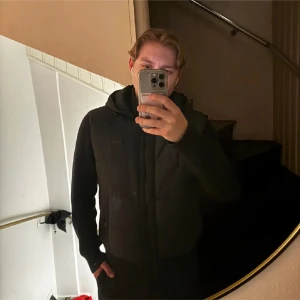 Svart jacka med huva från Boss - Snygg svart jacka från Boss med huva och dragkedja. Jackan har stickade ärmar och ribbade muddar, vilket ger en cool kontrast till det quiltade materialet på kroppen. Perfekt för kyligare dagar.