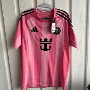 Rosa fotbollströja från Adidas - Säljer en rosa fotbollströja från Adidas med svarta detaljer och ränder på axlarna. Tröjan har korta ärmar och ett tryck med nummer 10 på baksidan. Perfekt för fotbollsfans som vill sticka ut på planen!