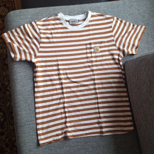 Brun och vit randig t-shirt från Carhartt - Säljer en snygg brun och vit randig t-shirt från Carhartt. T-shirten har en bröstficka med Carhartt-logga och är kortärmad. Perfekt för en avslappnad stil.