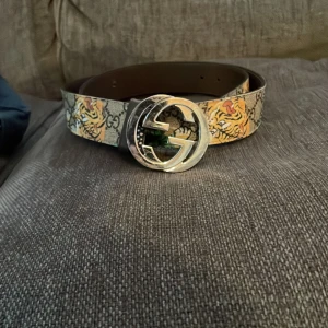 Mönstrat bälte med tiger och GG-logga - Snyggt limiterat bälte från Gucci med ett djärvt tiger- och lövmönster på en beige bakgrund. Bältet har en stor, rund spänne med den ikoniska GG-loggan i silver. Storlek 105. Nypris cirka 5000kr.