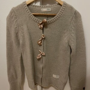 Odd Molly Beige stickad koft  - Charmig beige stickad kofta från Odd Molly med dekorativa rosetter framtill. Koftan har långa ärmar och ribbade kanter. Perfekt för kyliga dagar och ger en söt touch till din outfit. St 2