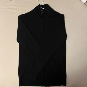 Merino full-zip från Oscar Jacobson -  Marinblå full-zip från Oscar Jacobson gjord i 100% merinoull. Perfekt för en stilren look. Passar  Tröjan har ett väldigt litet hål på framsidan som knappt syns nu mera då jag har lagat det, (bild 2 & 3) men är annars som ny.                 Nypris ca:2000 