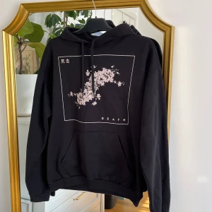 Svart hoodie med tryck från B&C Collection - Säljer en svart hoodie från B&C Collection med ett stilrent blommönster i vitt/ljusrosa och texten 'DEATH'. Hoodien har en klassisk passform med känguruficka och justerbar huva. Perfekt för en avslappnad stil.
