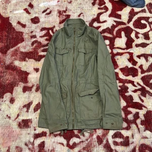 Grön jacka från H&M Divided - Säljer en grön jacka från H&M Divided med dragkedja och knappar. Jackan har långa ärmar och flera fickor framtill. Perfekt för höst och vår med sin stilrena design och praktiska detaljer.