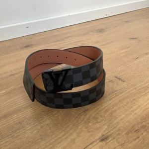 Svart och brun bälte med rutigt mönster - Snyggt bälte från  med ett klassiskt rutigt mönster i svart och brunt. Bältet har en stilren spänne med märkets logotyp. Perfekt för att ge en lyxig touch till din outfit.