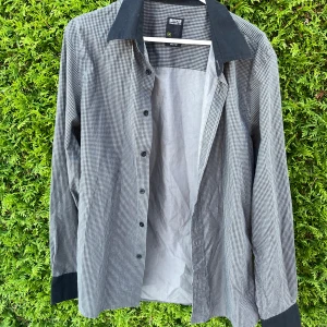 Rutig skjorta med svart krage - Snygg rutig skjorta i svart och vitt med kontrasterande svart krage och svarta muddar. Skjortan har klassisk passform, knappar framtill och lång ärm. 