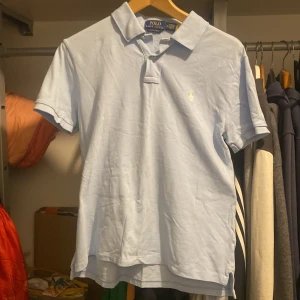 Ljusblå pikétröja från Polo Ralph Lauren - Snygg ljusblå pikétröja från Polo Ralph Lauren med klassisk krage och korta ärmar. Tröjan har den ikoniska broderade loggan på bröstet och är gjord i mjuk bomull. Perfekt för en clean och stilren look.