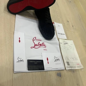 Christian Louboutin Svarta skor  - Snygga svarta sneakers från Christian Louboutin med klassiska röda sulor och coola svarta nitar på tån. Skorna har snörning och är tillverkade i skinn för en lyxig känsla. Perfekta för dig som vill sticka ut med exklusiva detaljer.