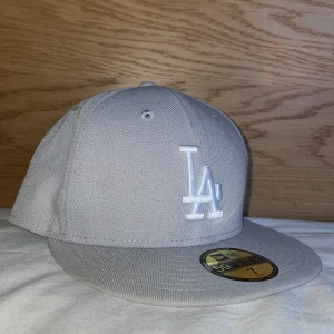 Grå New Era 59FIFTY LA keps - Snygg grå New Era 59FIFTY keps med LA-logga broderad framtill i vitt. Klassisk flat peak och strukturerad passform. Tillverkad i 100% polyester och perfekt för dig som gillar streetwear och sportig stil. Använd en gång