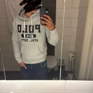 Grå hoodie från Polo Ralph Lauren - Säljer en grå hoodie från Polo Ralph Lauren med svart tryck framtill: 'POLO 1967 ATHL. DEPT.'. Tröjan har huva med dragsko och en stor magficka. Perfekt för en avslappnad stil.