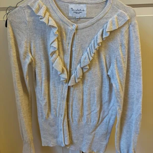 Ljusgrå/beige kofta med volang från Bondelid - Supersöt ljusgrå/beige kofta från Bondelid med volangdetaljer längs knäppningen och rund hals. Långärmad och stickad i en mjuk mix av viskos, polyamid och ull. Broderad patch med röd-vit hjärtdetalj på ärmen. Perfekt för lager-på-lager och chill vibes.