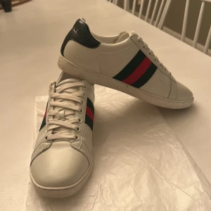 Vita sneakers från Gucci  - Snygga vita sneakers från Gucci med klassiska gröna och röda ränder på sidan. Skorna har snörning och en unik detalj i form av en  mörkgrön, ormskinnsmönstrad häl. Perfekt för en stilren look. Använda fåtalet gånger 