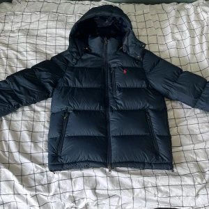 Mörkblå dunjacka från Polo Ralph Lauren - Snygg mörkblå dunjacka från Polo Ralph Lauren med huva, dragkedja och två sidofickor. Jackan har det klassiska röda Polo-loggan broderad på bröstet och justerbar huva med snörning. Perfekt för kalla dagar och riktigt stilren look. Priset är inte hugget i sten och skriv för fler bilder/ funderingar.
